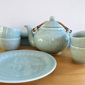 Może przedstawiać: Zestaw do herbaty w kolorze celadonu, składający się z czajniczka z plecionym uchwytem, półmiska i czterech filiżanek. Czajniczek i filiżanki mają wzór ryby. Zestaw jest ceramiczny i stoi na drewnianej powierzchni.