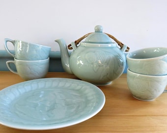 May include: A celadon tea set comprising a teapot with a woven handle, a serving platter, and four teacups. The teapot and teacups feature a fish design. The set is ceramic and is displayed on a wooden surface.