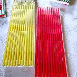 Könnte beinhalten: Zwei Packungen konischer Kerzen in transparenten Kunststoffbehältern. Eine Packung enthält gelbe Kerzen, die andere rote Kerzen. Die Kerzen sind in ordentlichen Reihen angeordnet, die Spitzen zeigen nach oben. Die Verpackung umfasst grün-weiß gestreifte Schachteln.