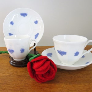 Peut inclure: Tasses et soucoupes blanches avec des motifs floraux bleus. Une tasse est sur un support en bois. Une rose en feutre rouge avec une tige verte est au premier plan. Sur une table en bois.