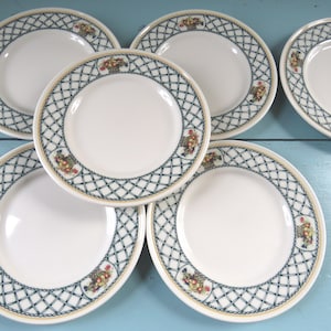 Puede incluir: Un juego de platos de cerámica blanca con un borde decorativo. El borde presenta un patrón de celosía azul y blanco con pequeños motivos de cestas florales. Los platos tienen un borde de color crema.