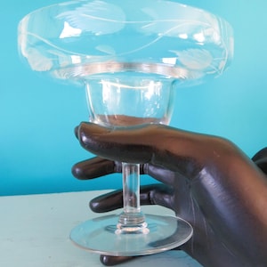 May include: Clear glass margarita glass with an etched leaf design around the rim. The glass has a wide bowl and a slender stem, ideal for cocktails. The glass is held by a black hand.