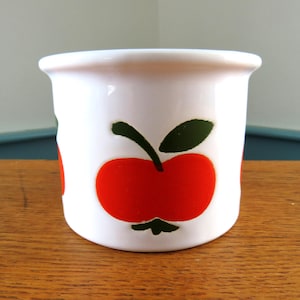 May include: A white ceramic mug featuring a red apple design with green leaves and stem. The cylindrical mug has a flared rim. The mug is displayed on a wooden surface. The design is repeated around the mug.
