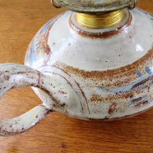 May include: A ceramic oil lamp with a round, off-white body and a large handle. The lamp features brown and blue painted accents. The top has a brass-colored metal fitting and a glass globe. A unique, handcrafted home decor item.