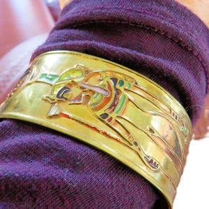 May include: A gold-colored cuff bracelet with an Egyptian-style design. The bracelet features a detailed relief with colorful accents of red, green, and brown. The cuff is worn on a purple sleeve.