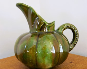 May include: A green and brown ceramic pitcher with a bulbous body and a curved spout. The handle is adorned with small, raised dots. The pitcher is on a wooden surface.