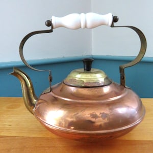 May include: A copper tea kettle featuring a rounded body, a curved brass spout, and a brass lid topped with a small black knob. The kettle has a high, arched metal handle fitted with a smooth, white, cylindrical grip in the centre.