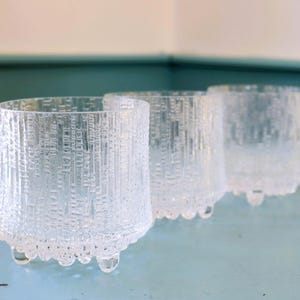 May include: Three clear, textured glass tumblers with a cylindrical shape and small, rounded feet. The glasses have a textured, vertical pattern and are arranged in a row, showcasing their unique design.