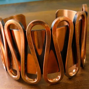 May include: A copper-toned cuff bracelet with a unique design. The bracelet features a series of curved, elongated loops connected by a dark, contrasting interior. The metal has a warm, polished finish, creating a striking visual effect. This statement piece is a bold accessory.