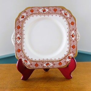 May include: Square white plate with a decorative border featuring orange floral and geometric patterns, and gold trim. The plate is displayed on a red stand, showcasing its design and shape. A vintage piece for serving or display.