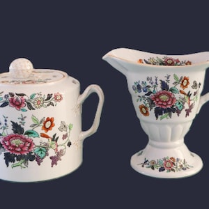 May include: A white ceramic sugar bowl with a lid and a creamer pitcher, both adorned with a floral pattern in shades of pink, orange, green, and blue. The sugar bowl has two handles and the creamer has a handle and a spout.