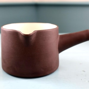 May include: A small, brown ceramic jug with a handle and a spout. The interior is cream-coloured. The jug is on a light blue surface. It is likely used for serving sauces or liquids.