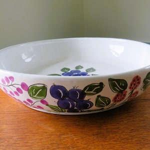 Puede incluir: Bol de cerámica blanco con un diseño floral pintado a mano. El cuenco presenta ilustraciones botánicas moradas, rosas y verdes alrededor del borde. Una pieza decorativa para servir ensaladas o exhibir fruta.