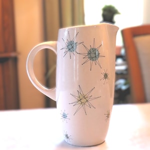May include: A cream-colored ceramic pitcher featuring a handle and a spout, decorated with a repeating pattern of black starburst lines overlaid on small, pale blue and light yellow circular shapes scattered across the exterior surface.