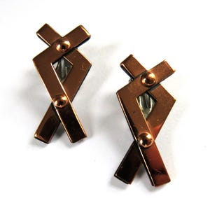 May include: A pair of copper-colored clip-on earrings. Each earring features an abstract geometric design with intersecting rectangular bars and small, round rivets. The earrings have a shiny, metallic finish.