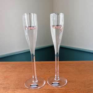 May include: Two clear glass candlestick holders with a faceted design. Each has a round base and a slender stem that widens to hold a candle. The brand name "ONEIDA" is visible on the base of each holder. The background is a wooden surface.