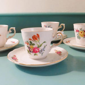 May include: Four white teacups and saucers with floral designs. Each cup and saucer has a gold trim. The teacups feature different floral arrangements in shades of pink, yellow, and purple. The saucers have a scalloped edge.