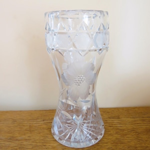 May include: A clear glass vase with a flared base and a wide opening. The vase features etched floral and geometric patterns, including a large flower design in the center. The vase is displayed on a wooden surface.