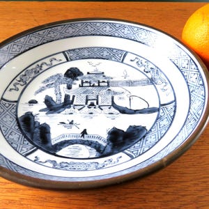 May include: A decorative ceramic plate with a blue and white Asian-inspired landscape design. The plate has a brown rim and is next to a whole orange fruit on a wooden surface.