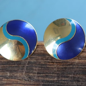 May include: A pair of round stud earrings. Each earring features a gold-colored base with a curved section of vibrant blue enamel and a turquoise enamel accent. The earrings are displayed on a wooden surface.