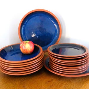 May include: A set of blue ceramic plates with a terracotta-colored rim. The plates are stacked in several piles, with one plate on top of a stack holding a red and yellow apple. The plates are arranged on a wooden surface.