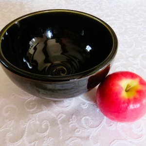 May include: A dark brown ceramic bowl with a glossy finish, next to a red apple. The bowl has a spiral design at the bottom. The apple is red with yellow accents.