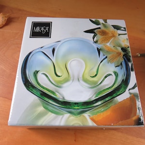 May include: A Mikasa glass bowl in its original box. The bowl has a unique shape with green, yellow, and blue hues. The box features the Mikasa logo and floral imagery. A decorative piece for the home.