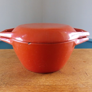 May include: A small, orange cast iron pot with a lid. The pot has two handles and sits on a wooden surface. The lid is flat and fits snugly on top of the pot. The pot is a vibrant shade of orange.