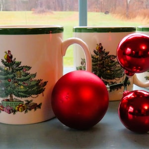 Op de afbeelding: Vier witte keramische mokken met een groene rand en een kerstboomontwerp, samen met drie rode kerstballen. De mokken hebben een gedetailleerde illustratie van een versierde kerstboom.