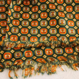 Op de afbeelding: Een groene sjaal met een ingewikkeld patroon van oranje, blauwe en gele vierkanten. De sjaal heeft een gefranjerde rand, wat textuur en detail toevoegt. Het ontwerp herhaalt zich over de stof, waardoor een visueel aantrekkelijk accessoire ontstaat.