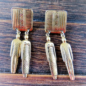 May include: A pair of gold-coloured earrings. Each earring has a rectangular top with a geometric design and a dangling figure. The figures have etched details, creating a vintage or tribal aesthetic. The earrings are displayed against a wooden background.