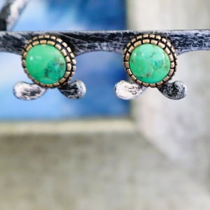 Peut inclure: Une paire de boucles d'oreilles à clous avec des pierres rondes d'un vert éclatant présentant des motifs naturels de matrice brune. Chaque pierre est sertie dans un chaton métallique texturé à bord perlé, sur un support sombre.