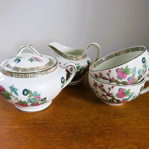 May include: A white porcelain tea set with floral and geometric patterns. The set includes a lidded sugar bowl, a creamer jug, and two teacups. The floral design features pink and blue flowers with green foliage.