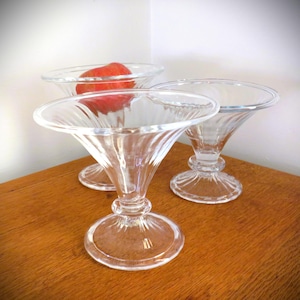 Pode incluir: Três taças de sobremesa de vidro transparente com uma tigela alargada e uma base de pedestal. Uma taça contém uma fatia de maçã vermelha. As taças estão dispostas sobre uma superfície de madeira, com uma parede branca ao fundo.