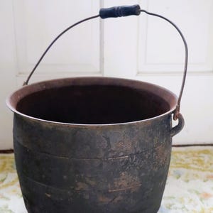 May include: A dark, weathered metal bucket with a rounded body and horizontal ridges. It features a thin, arched wire handle with a central black wooden grip. The exterior surface shows signs of oxidation and a textured, aged patina.