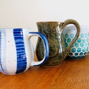 May include: Three ceramic mugs of varying designs and colors sit on a wooden surface. One mug is white with blue stripes, another is green with a textured pattern, and the third is blue and white with a honeycomb design.