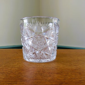 May include: A clear, cut-glass tumbler with a detailed starburst and geometric pattern. The glass has a cylindrical shape and sits on a wooden surface. The intricate design reflects light, creating a sparkling effect. This vintage glassware is perfect for serving beverages.