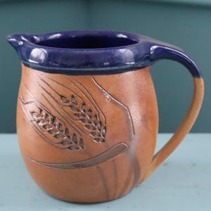 May include: A handcrafted ceramic pitcher with a brown body and a dark blue rim and handle. The pitcher features an embossed wheat stalk design. The handle is a lighter brown color. This decorative pitcher is suitable for serving beverages.
