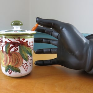 Könnte beinhalten: Zylindrisches Keramikgefäß mit Deckel, verziert mit handgemalten Pfirsichen und grünen Blättern auf weißem Grund. Der Deckel hat einen grünen Knopf. Dekoratives Gefäß für die Küche.