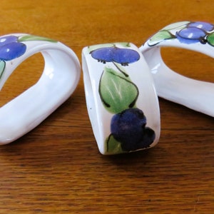 Op de afbeelding: Drie witte keramische servetringen met een handgeschilderd ontwerp van blauwe bessen en groene bladeren. De servetringen zijn ovaal van vorm en liggen op een houten oppervlak. Het ontwerp wordt op elke ring herhaald.