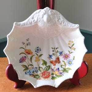 May include: White porcelain decorative dish with a scalloped edge and floral design. The dish features colorful flowers, including roses, and a butterfly. The dish is displayed on a red stand.