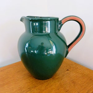 May include: A glossy, dark green ceramic pitcher with a rounded body and a curved handle. The handle has a terracotta-colored accent. The pitcher sits on a wooden surface, reflecting the light.