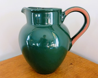 May include: A glossy, dark green ceramic pitcher with a rounded body and a curved handle. The handle has a terracotta-colored accent. The pitcher sits on a wooden surface, reflecting the light.