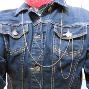 May include: Dark blue denim jacket with button closure, two chest pockets, and gold stitching. A gold chain necklace is draped over the jacket. The jacket has a classic collar and buttoned cuffs. A scarf is visible at the neckline.