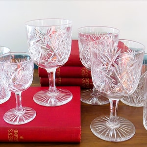 May include: A collection of clear crystal glassware, including wine glasses, goblets, and tumblers, with a starburst pattern. The glasses are arranged on a wooden surface and red books.