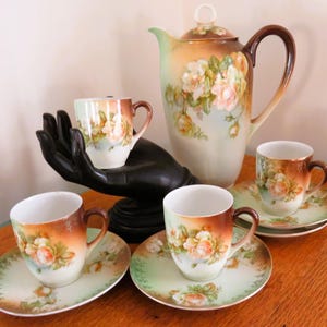 May include: A vintage tea set featuring a teapot, cups, and saucers. The set has a floral design with shades of orange, brown, and green. The cups and saucers are arranged on a wooden surface, with one cup held by a black hand sculpture.