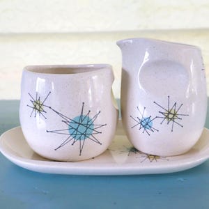 May include: A ceramic creamer and sugar bowl set on an oval tray, featuring a speckled off-white finish with black starburst patterns and light blue and yellow circular accents. The pieces have rounded shapes and a smooth, glazed surface texture.