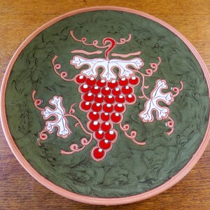 May include: A round ceramic plate with a green background and a red grape design. The grapes are surrounded by white leaves and vines. The rim of the plate is a light brown color.