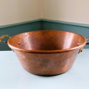 May include: A round copper basin with a polished finish and two curved brass handles attached to the sides with rivets. The bowl features a wide, open top and a smooth, slightly tapered base, showcasing the natural metallic texture of the copper.