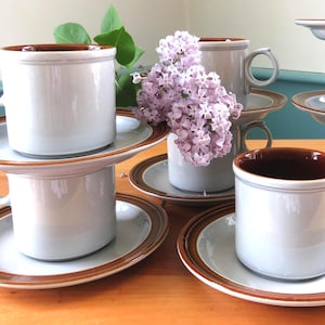 May include: A set of white ceramic mugs with brown interiors and matching saucers. The rims of the mugs and saucers have a brown border. A small bouquet of purple flowers sits in a mug. The mugs and saucers are arranged on a wooden surface.